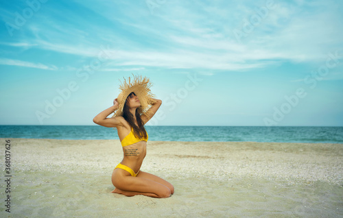 Beautiful tanned woman on tropical beach by ocean with copy space. girl wearing in yellow bikini and big straw hat sits on sand, breathing and enjoying sea breeze