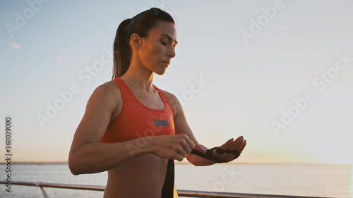 Young lady is wrapping her arm with black boxing wrist wrap. She standing near the seacoast during her training on embankment. Slow motion photo