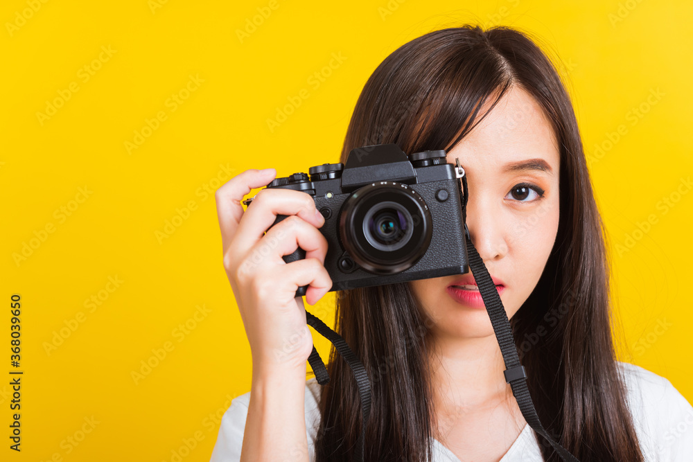 Portrait of happy Asian beautiful young woman photographer taking a picture and looking viewfinder on retro digital mirrorless photo camera ready to shoot, studio shot isolated on yellow background