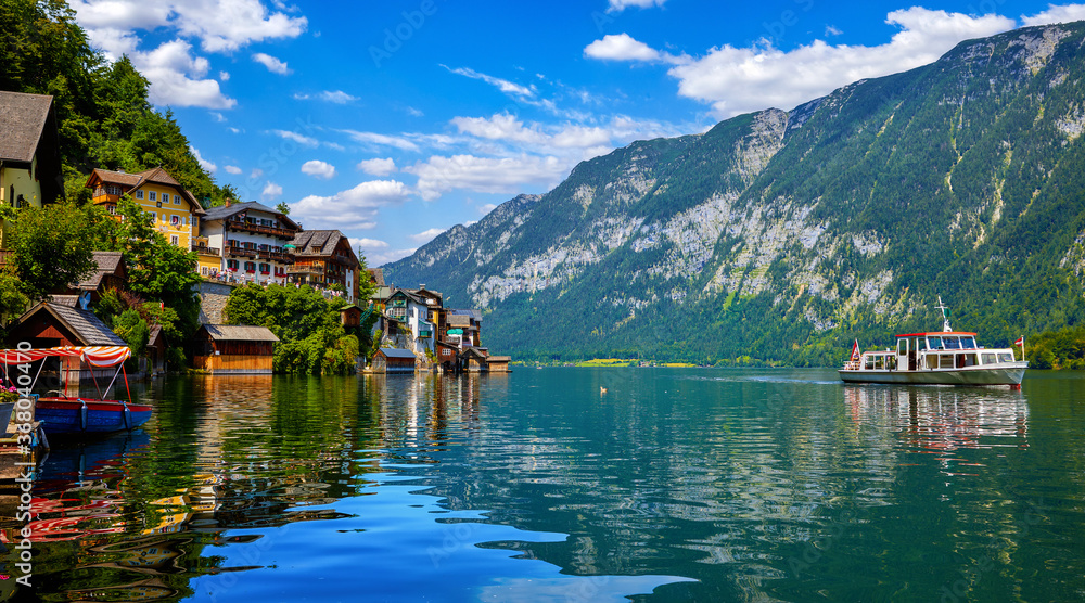 Hallstatt, Austria. Lakeside town in the Austrian Alps. Classic postcard view of famous landscape with passenger ship on a water. Beautiful sunny day with blue sky and clouds. Salzkammergut region - obrazy, fototapety, plakaty 