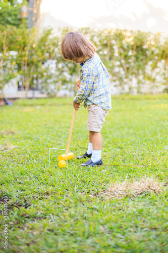 Child wearing bow tie and plaid button down playing croquet on grass yellow stick and ball