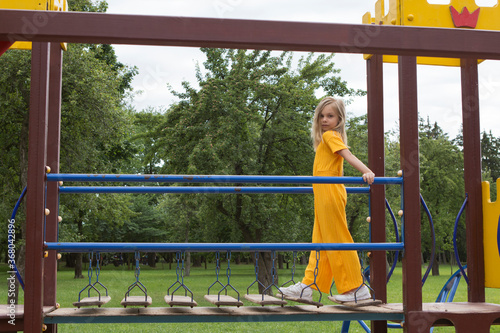 A beautiful six-seven-year-old girl with blond hair in a bright yellow jumpsuit walks across the bridge on a children's playground in summer.
