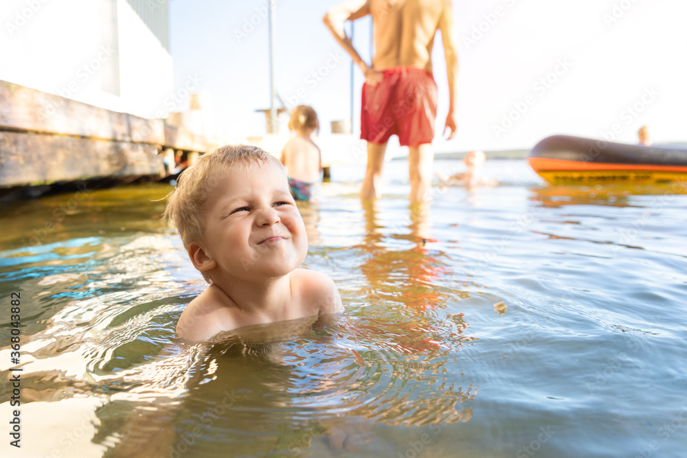 Cute adorable cheerful caucasian little blond toddler boy enjoy having fun playing at lake or river beach water on warm sunset evening time outdoors.. Happy childhood vacation at countryside concept