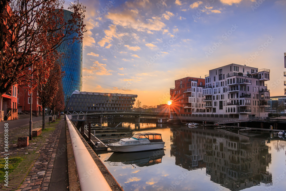 Morning mood in a residential area. Sunrise with sun rays between the buildings. River course in the west port of the city of Frankfurt. Street with trees, bench and boat in the harbor