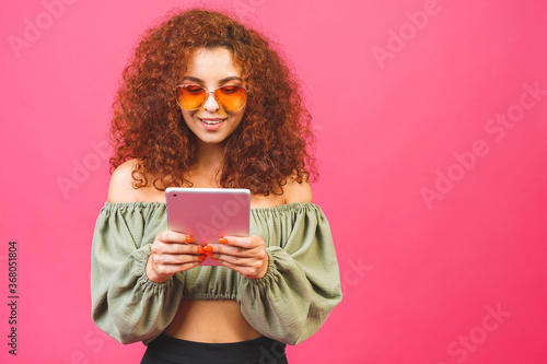 A young girl in sunglasses and holding a tablet computer and smiling. Portrait of a girl isolated over pink background with space for text.