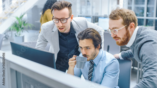 In Bright Modern Office: Young Businessman Sitting and Working at His Desktop Computer with Project Manager and Team Leader Standing Beside Him, Have Discussion, Finding Problem Solution 