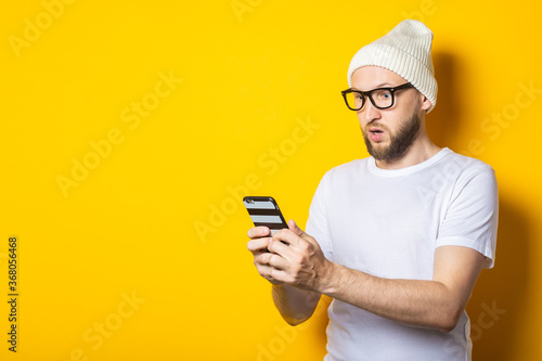 Bearded young man in a hat and glasses looks surprised in surprise at the phone on a yellow background. © Alex