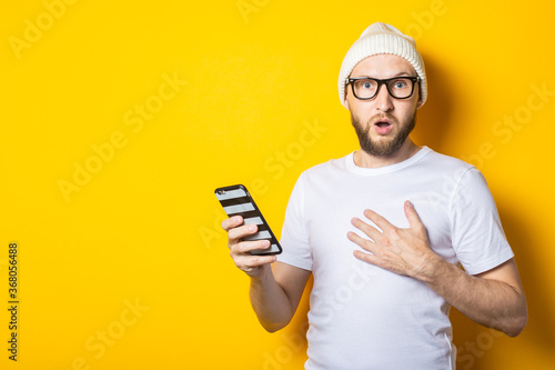Bearded young man in a hat and glasses holding a phone in surprise on a yellow background.