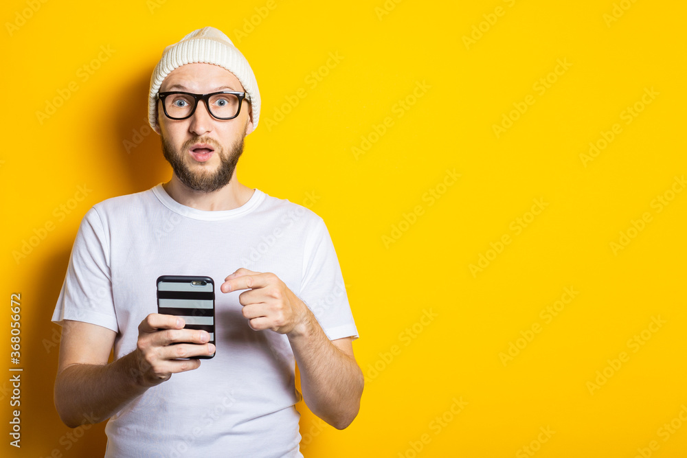 Surprised young man with a beard points his finger at the phone on a yellow background Surprised young man with a beard points his finger at the phone on a yellow background