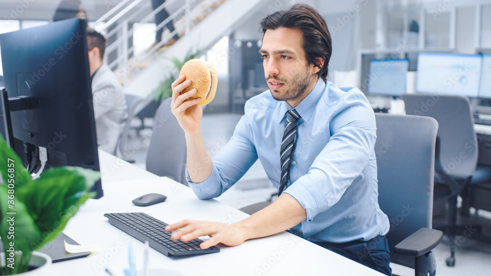 In the Bright Modern Office Young Businessman Eats Hamburger Sitting at His Desktop Computer, Prepared to Bite it and Continue to Work During His Lunch. In the Background Colleagues Working In the Bright Modern Office Young Businessman Eats Hamburger Sitting at His Desktop Computer, Prepared to Bite it and Continue to Work During His Lunch. In the Background Colleagues Working