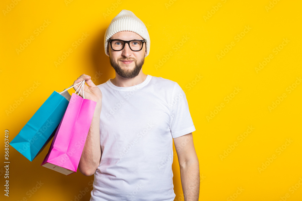 Friendly smiling young man with a beard, in glasses and a hat, holds packages with purchases on a yellow background.