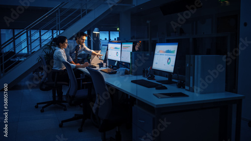 Late at Night In Modern Office: Businessman and Businesswoman Work on Desktop Computer, Having Discussion, Finding Problem Solution, Finishing Project. Successful Dedicated Responsible Office Workers