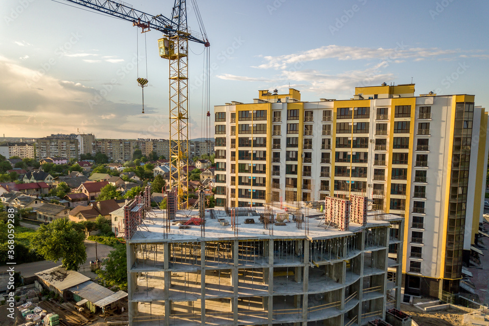 Apartment or office tall building under construction. Brick walls, glass windows, scaffolding and concrete support pillars. Tower crane on bright blue sky copy space background. Apartment or office tall building under construction. Brick walls, glass windows, scaffolding and concrete support pillars. Tower crane on bright blue sky copy space background.