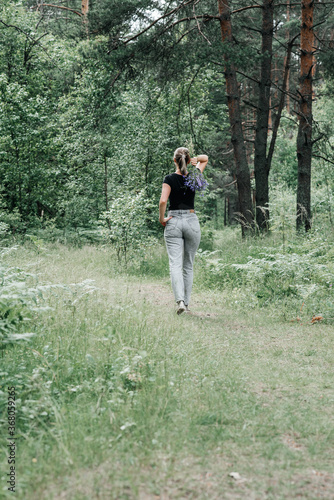 young woman with a tail of hair in a black t-shirt and gray jeans in the woods holding blue wildflowers in her hands © Виктория Бычкова