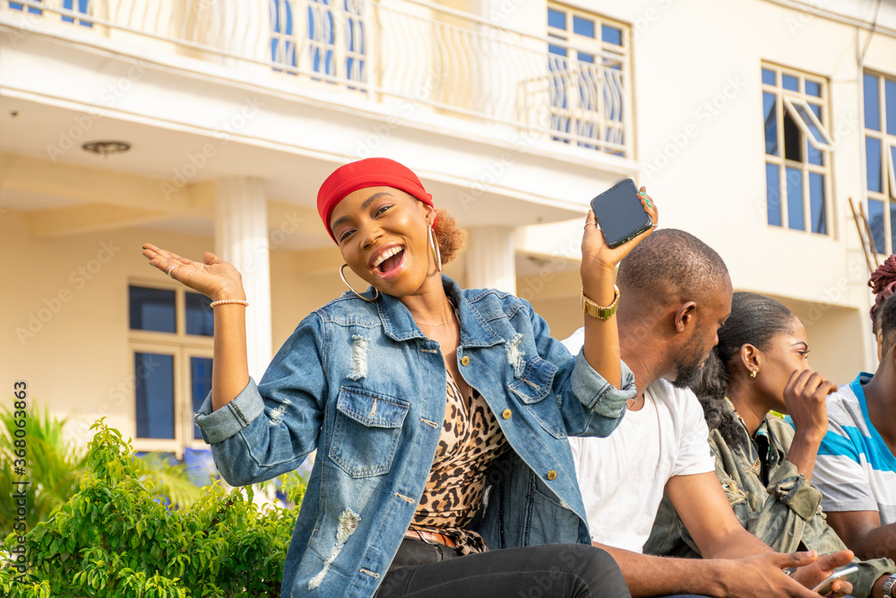 an excited young black woman sitting with her friends, holding her phone an excited young black woman sitting with her friends, holding her phone