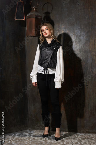 Fashion vogue style full length portrait of young gorgeous blonde female model in black trousers and white blouse standing against grunge metal wall