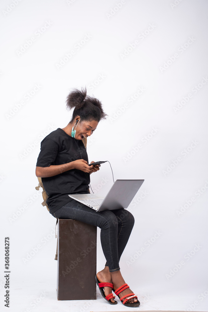 african girl wearing a face mask, carrying a backpack, sitting and using her mobile phone and laptop, looking surprised