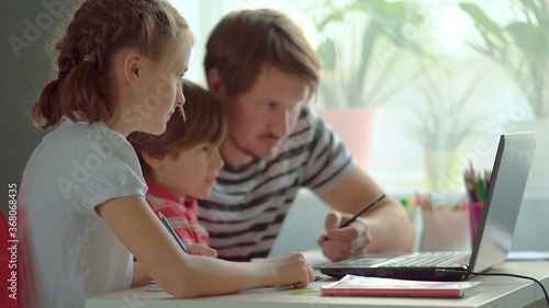 Cute children use laptop for education, online study, home studying, Boy and Girl have homework at distance learning. Lifestyle concept for home schooling. Father helps daughter and son.