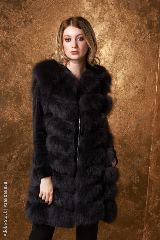 Young beautidul glamorous woman in black luxury expensive fur waist coat posing against golden wall. Fashion portrait
