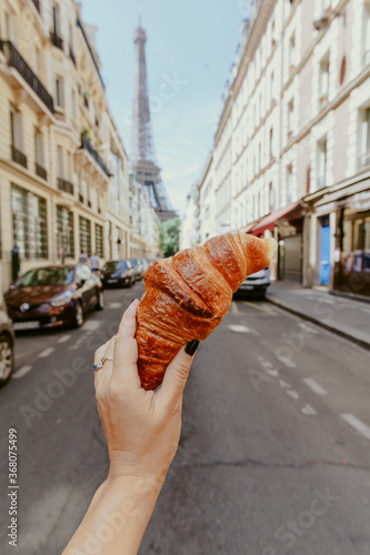 Photography Hand holding croissant in parisian street with Eiffel tower view