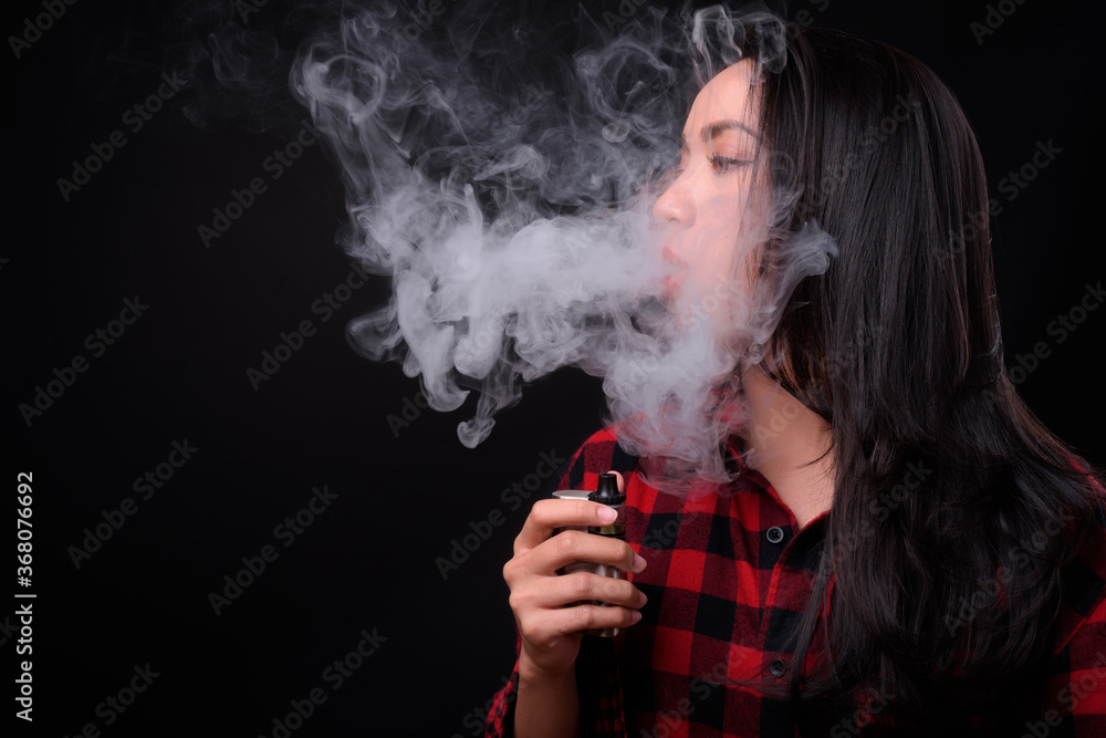 Young beautiful Asian woman vaping against black background Young beautiful Asian woman vaping against black background