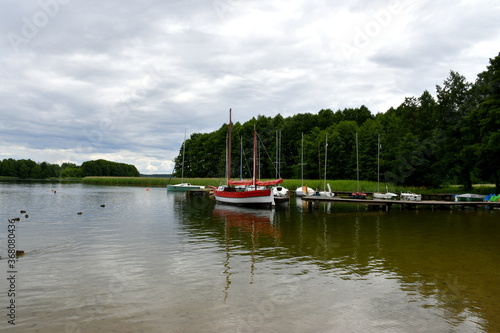 A view of a vast yet shallow lake with several boats parked next to its coast with a dense forest visible in the distance under the cloudy summer sky seen on Polish countryside during a hike