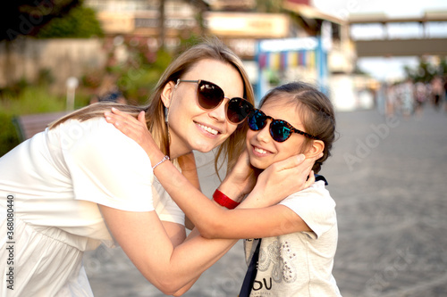 Outdoor portrait of beautiful blonde mother and her cute child. Small girl hugging her mom on the beach. Little lady and mom wearing sunglasses. They smiling. Summer sunny day. Happy Mothers day.
