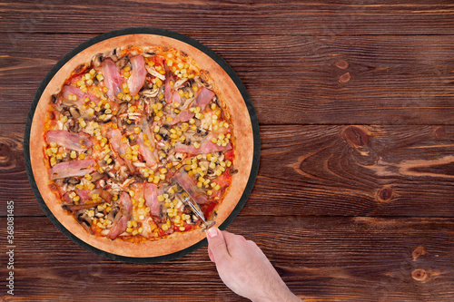Man cuts the tasty pizza with chicken breast, corn, bacon and mushrooms, with a pizza cutter on a black round plate which is on wooden rustic table, copy space