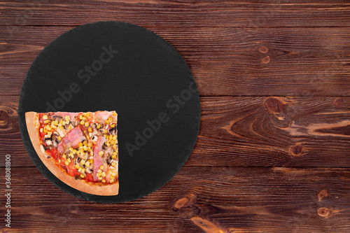 A quarter of italian pizza with chicken breast, corn, bacon and mushrooms, on a slate round plate which is on wooden background, top view and copy space