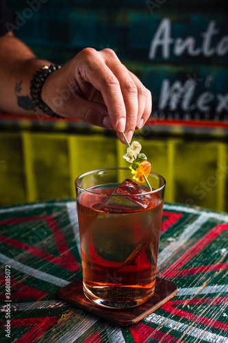 A transparent cocktail in a highball glass with a big ice cube. A hand putting gummy bears garnish on top of a drink