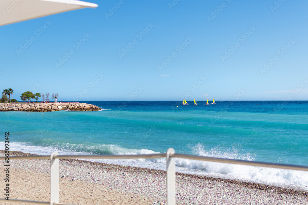 View from a cafe overlooking the beach and Mediterranean Sea with sailboats in the water in Menton, France, on the French Riviera.