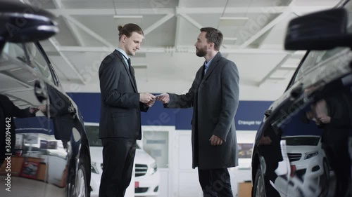 Adult man is giving money to manager and shaking hands with auto deaker in office in showroom. Handsome man is talking to the car dealer