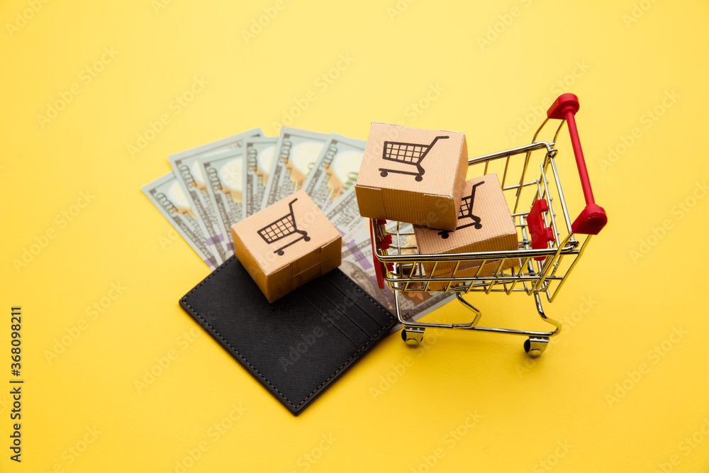 Grey wallet with hundred dollar bills, three delivery boxes and shopping trolley on yellow background. Grey wallet with hundred dollar bills, three delivery boxes and shopping trolley on yellow background.
