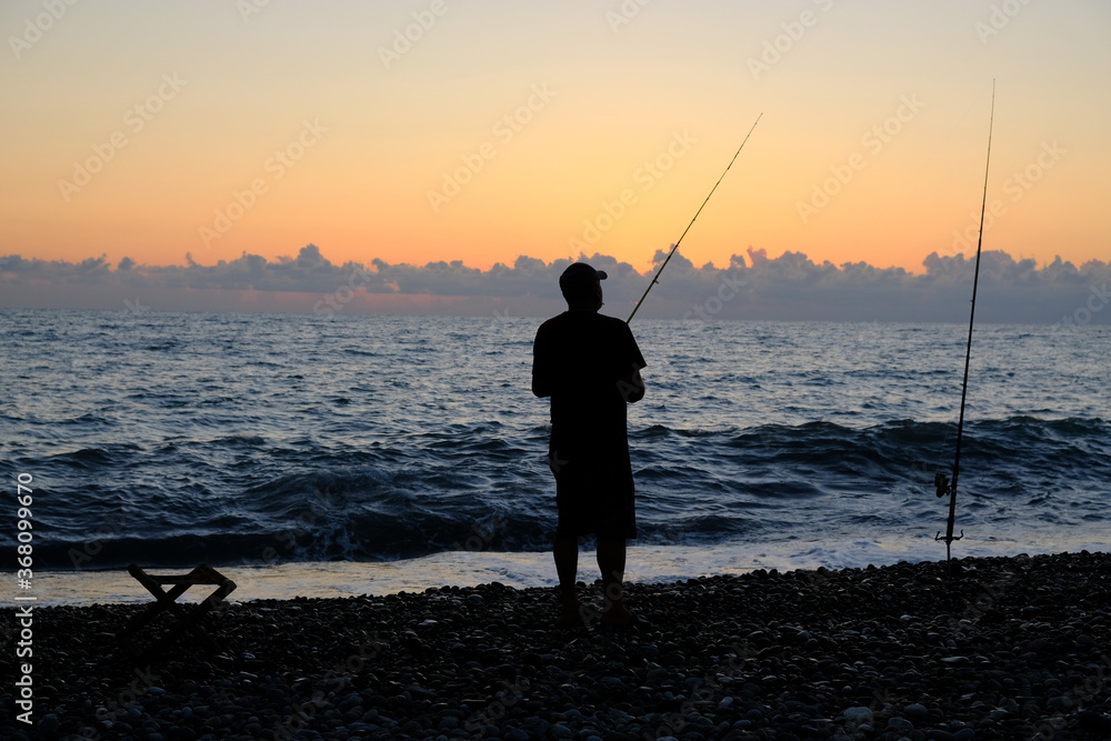 Silhouette of a fisherman against the background of the evening sky. Sunset at the sea. The man is holding a fishing rod. Summer. Georgia. Black Sea.