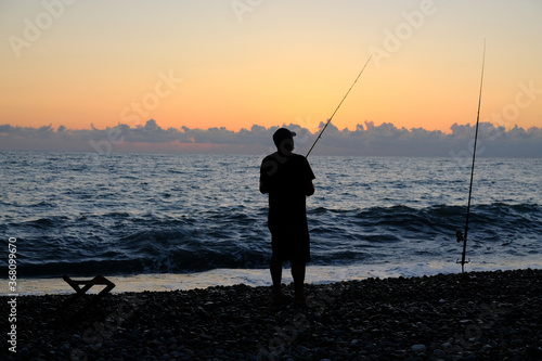 Silhouette of a fisherman against the background of the evening sky. Sunset at the sea. The man is holding a fishing rod. Summer. Georgia. Black Sea. © Viktor