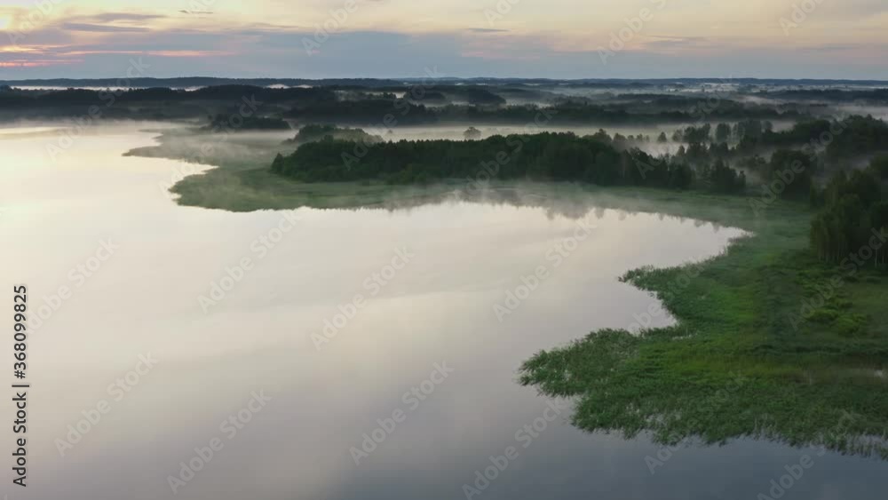 Aerial foggy morning at lake just before dawn. Drone flies over water surface and shore. Clouds of fog moving above ground. Coniferous forests and meadows on horizon. Idyllic landscape on sunrise Aerial foggy morning at lake just before dawn. Drone flies over water surface and shore. Clouds of fog moving above ground. Coniferous forests and meadows on horizon. Idyllic landscape on sunrise