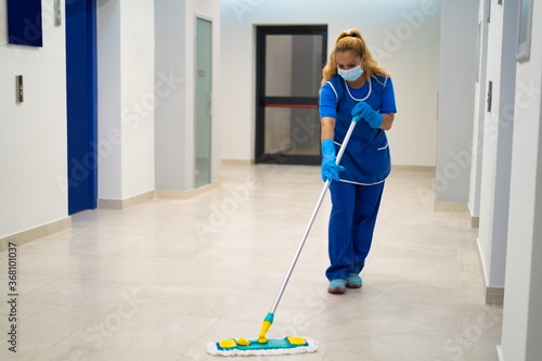 A cleaning lady with the mask on her face mops the floor in an office building photo