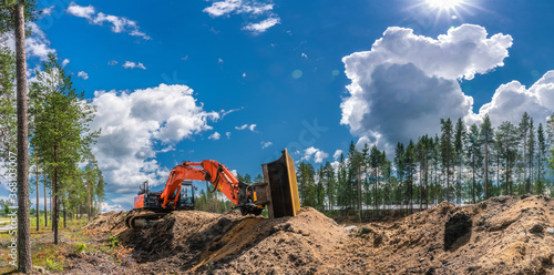 Panorama view on orange excavator that stands on top of digged from trench sand. Fully streched arm with bucket lies along the dirt close to the trench. Sunny day - clear blue sky and white clouds