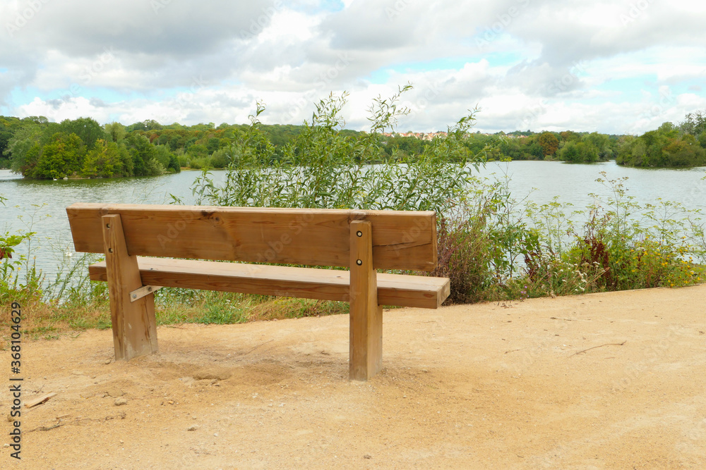 Rural landscape by the water. Isolated bench on the sand, facing a lake with the forest in the background on the horizon. Rural landscape by the water. Isolated bench on the sand, facing a lake with the forest in the background on the horizon.