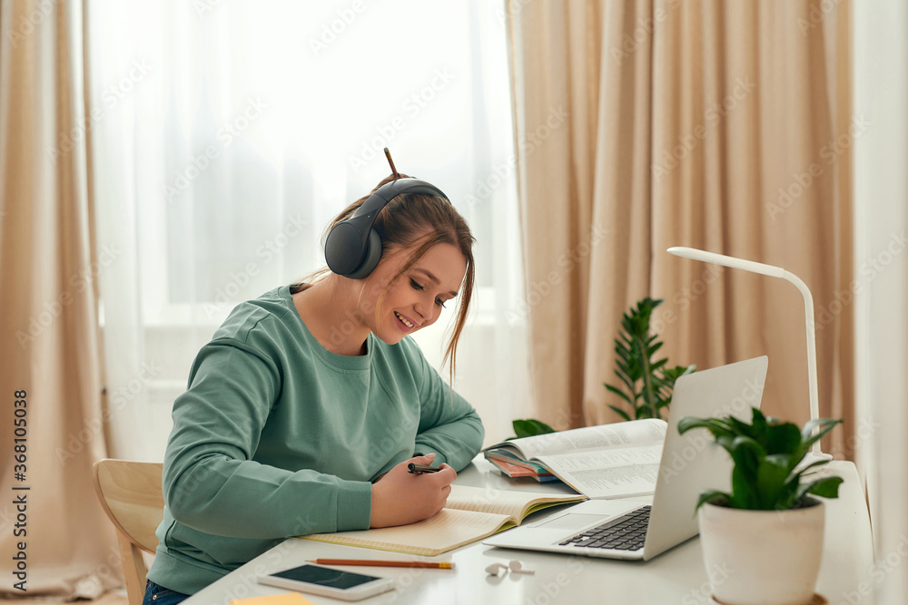 Studying online. Young beautiful woman, happy female student wearing headphones, listening online course and making notes while sitting at desk Studying online. Young beautiful woman, happy female student wearing headphones, listening online course and making notes while sitting at desk