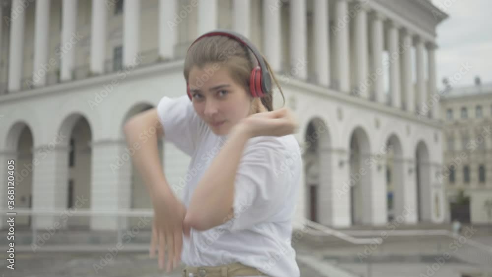 Middle shot of charming girl in headphones dancing robot dance with mouth closed with hand. Portrait of cheerful Caucasian young woman performing in city center on square. Modern arts, lifestyle. Middle shot of charming girl in headphones dancing robot dance with mouth closed with hand. Portrait of cheerful Caucasian young woman performing in city center on square. Modern arts, lifestyle.