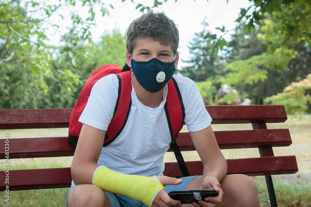 Fototapeta premium Close up young boy with protective mask on face on bench with phone. Teenager in white t shirt wearing medical mask to protect from corona virus, illness, infection. Quarantine on the city streets