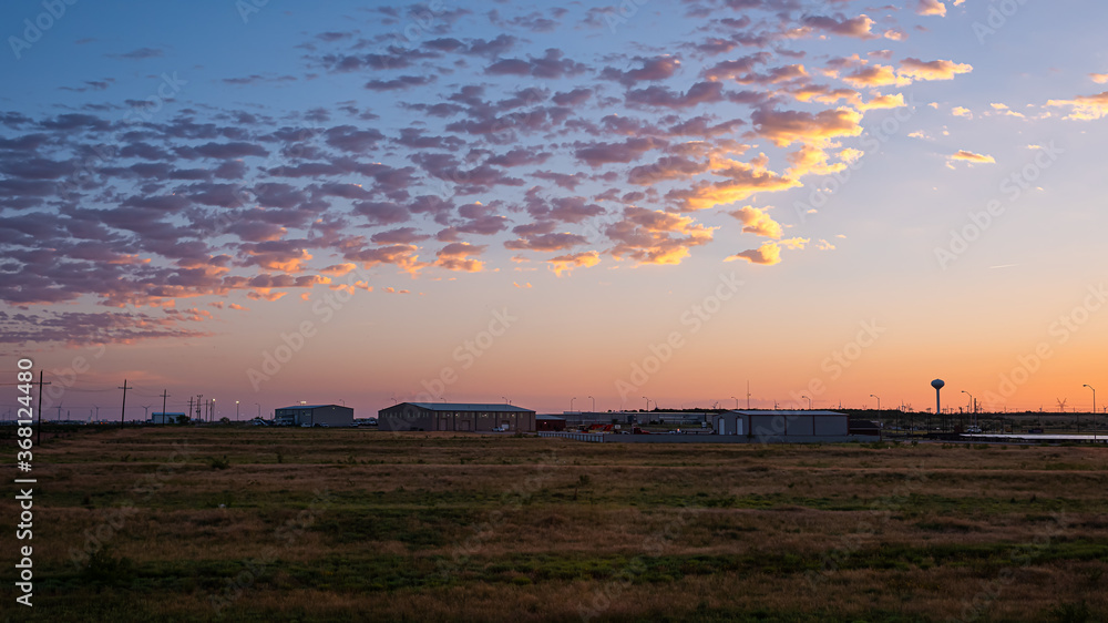 Fototapeta premium Colorful dark dawn sunrise in Snyder Texas with industrial building, warehouses or storehouses and factory by farm field with electricity pylons