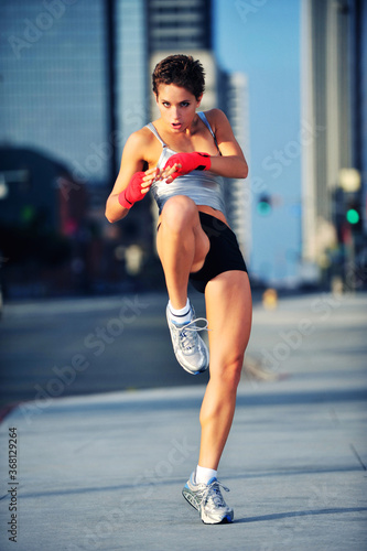 Pretty mixed martial arts young woman wearing boxing wraps and kicking with Los Angeles skyscrapers towering behind © Eric Hood