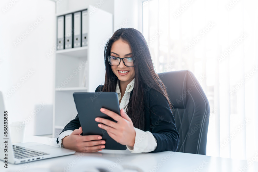 Cute female with long hair and glasses sitting in office and looking at tablet. Cute female with long hair and glasses sitting in office and looking at tablet.