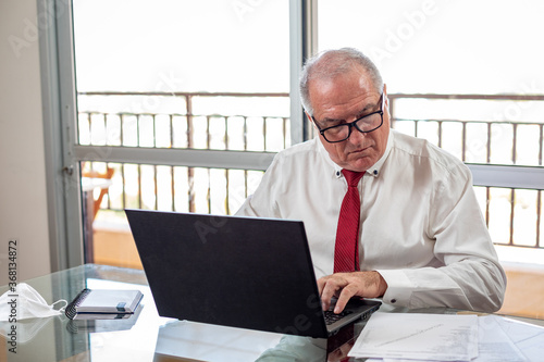 Sir typing on the notebook, working on the home office system in times of pandemic by the Corona virus. Mister works at home wearing a shirt and tie with his mask resting beside.