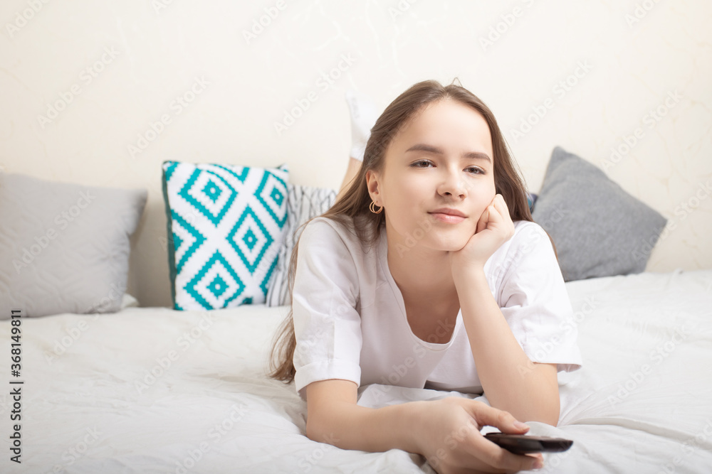 Home rest. Girl is lying at home on the bed with a remote control in her hands, watching tv, smiling Home rest. Girl is lying at home on the bed with a remote control in her hands, watching tv, smiling