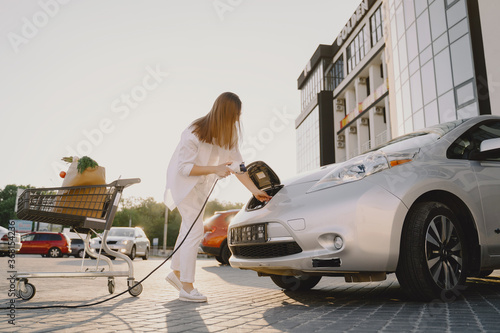 Charging electro car at the electric gas station. Woman standing by the car. Lady with foodstuff.