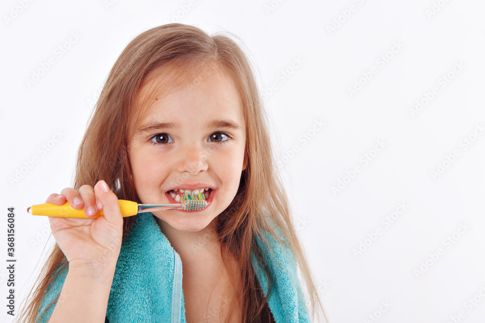 a little girl brushes her teeth on a white background. portrait of a child with a yellow toothbrush. blue towel around her neck. morning hygiene procedures a little girl brushes her teeth on a white background. portrait of a child with a yellow toothbrush. blue towel around her neck. morning hygiene procedures