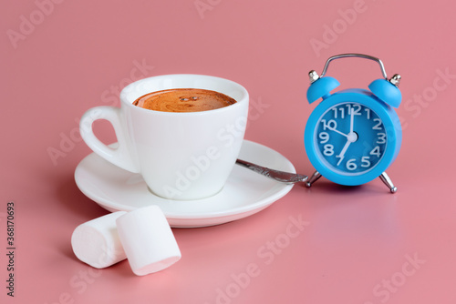 Morning cup of coffee, two marshmallows and blue alarm clock on pink background. 7 o'clock on clock face. Start of the day concept. Closeup. Copy space
